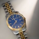 A luxurious Mathey-Tissot watch featuring a blue dial, gold-tone bezel, and a two-tone bracelet, illuminated by soft lighting.