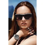 A person with long brown hair wears multiple rings and a stylish black watch while posing with a scenic background.