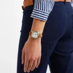 A close-up of a person's wrist wearing a silver watch with a brown leather strap, paired with navy pants and a striped blue shirt.