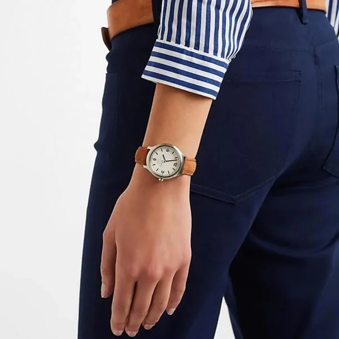 A close-up of a person's wrist wearing a silver watch with a brown leather strap, paired with navy pants and a striped blue shirt.