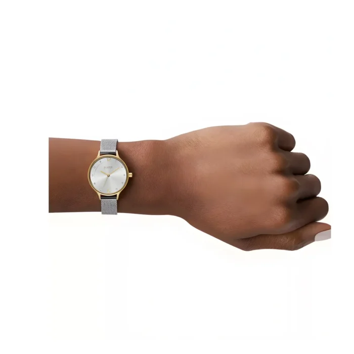 A close-up of a hand wearing a silver mesh band watch with a gold case and white face, resting on a light background.