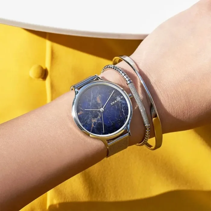 A close-up of a wrist wearing a Skagen watch with a deep blue marble face, paired with two sleek silver bracelets, against a yellow backdrop.
