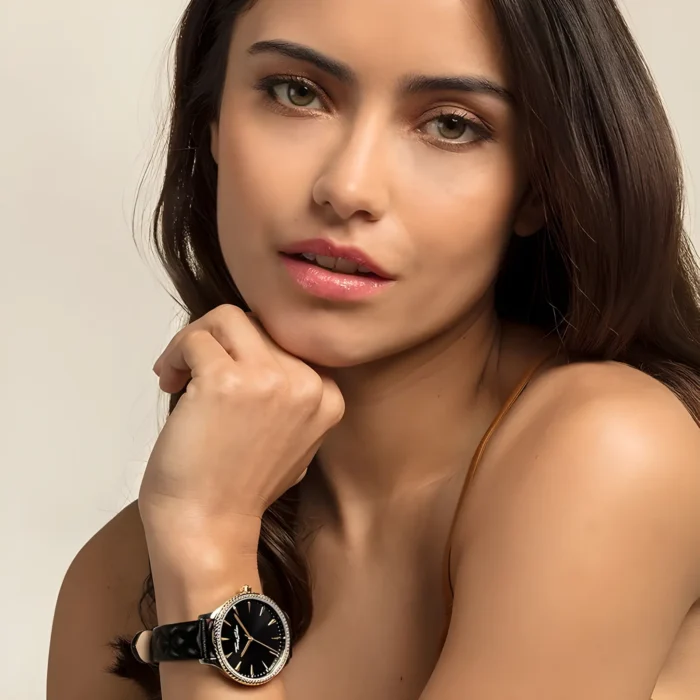 A close-up of a woman’s hand resting on her chin, showcasing an elegant black watch with a gold bezel, against a neutral background.