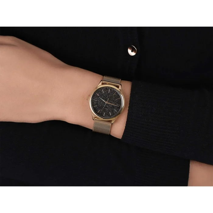 A close-up of a woman's hand wearing a stylish gold mesh wristwatch with a black textured dial against a dark sweater background.