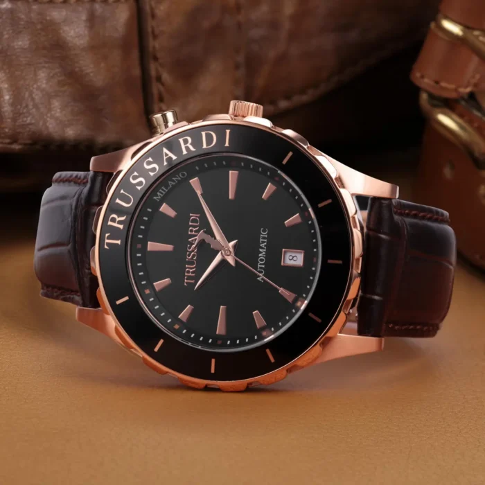 Stilingas „Trussardi“ automatinis laikrodis su juodu ciferblatu, rožinio aukso akcentais ir rudu odiniu dirželiu tekstūros fone.