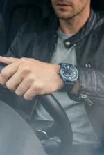 A man in a leather jacket, holding a steering wheel, wears a stylish watch with a black face and bold numbers, conveying a sense of sleek elegance.